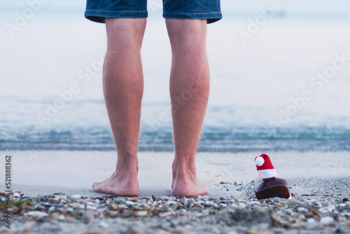 Christmas party, Xmas time. Single bottle of whisky with Santa hat in sand of the sea, legs of young man standing on the sand by sea. Festive decoration, soft focus. Seasonal holidays concept
