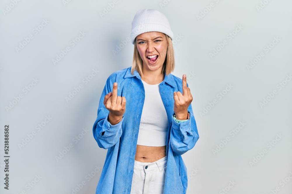 Beautiful blonde woman wearing wool hat showing middle finger doing fuck you bad expression, provocation and rude attitude. screaming excited