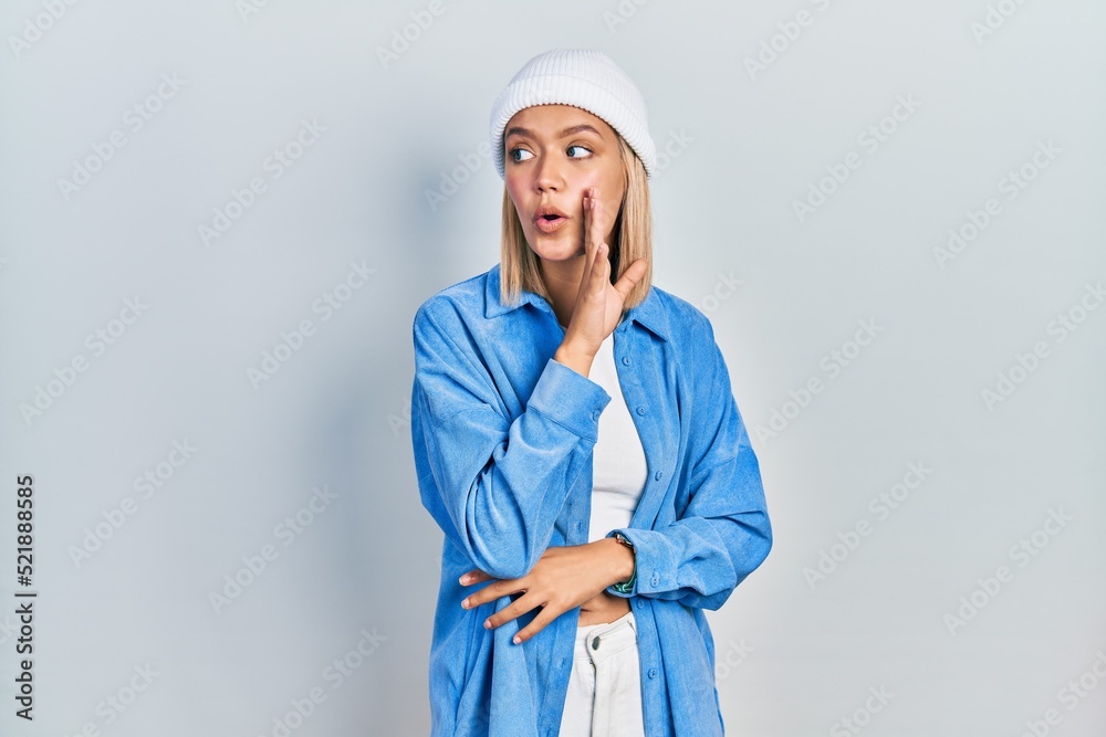 Beautiful blonde woman wearing wool hat hand on mouth telling secret rumor, whispering malicious talk conversation Beautiful blonde woman wearing wool hat hand on mouth telling secret rumor, whispering malicious talk conversation