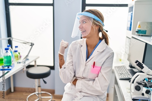 Young blonde woman working at scientist laboratory wearing face mask pointing thumb up to the side smiling happy with open mouth