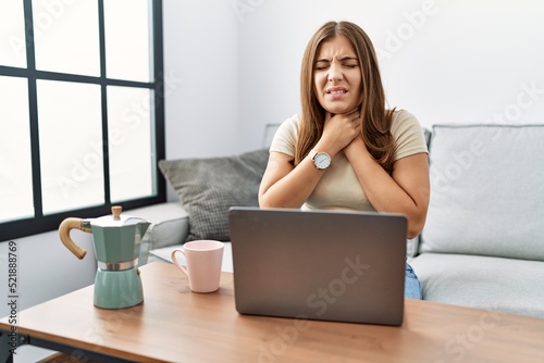 Young brunette woman using laptop at home drinking a cup of coffee shouting and suffocate because painful strangle. health problem. asphyxiate and suicide concept.