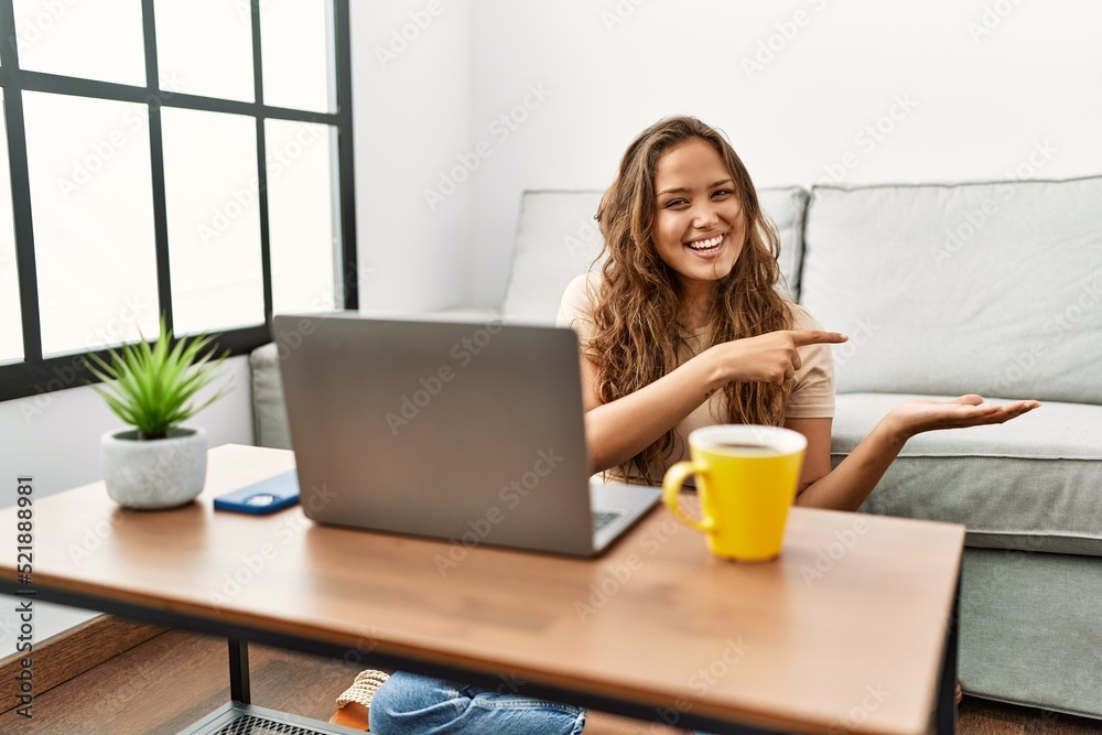 Beautiful hispanic woman using computer laptop at home amazed and smiling to the camera while presenting with hand and pointing with finger. Beautiful hispanic woman using computer laptop at home amazed and smiling to the camera while presenting with hand and pointing with finger.
