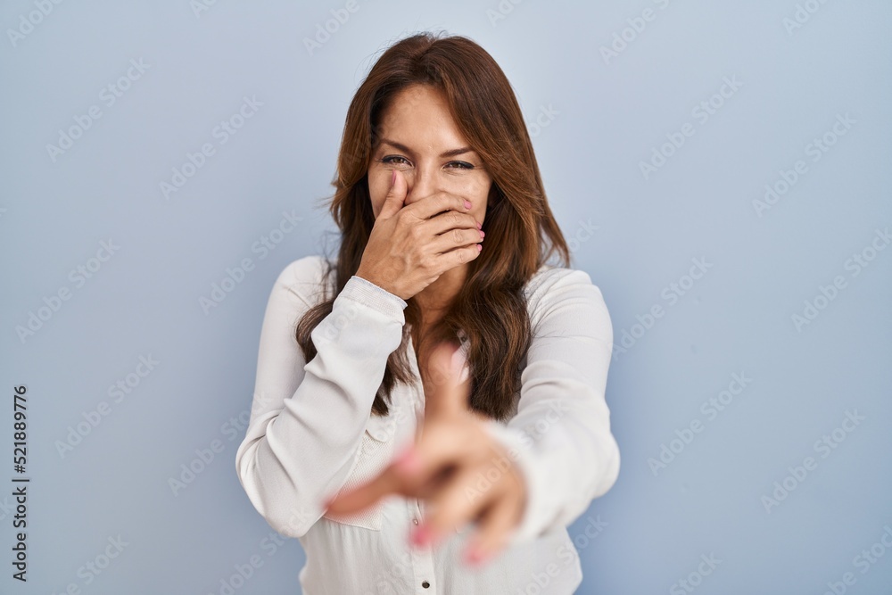 Hispanic woman standing over isolated background laughing at you, pointing finger to the camera with hand over mouth, shame expression Hispanic woman standing over isolated background laughing at you, pointing finger to the camera with hand over mouth, shame expression