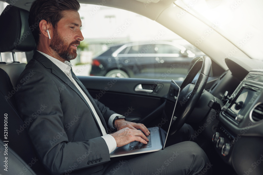 Handsome businessman working on laptop while sitting on driver seat in luxury car