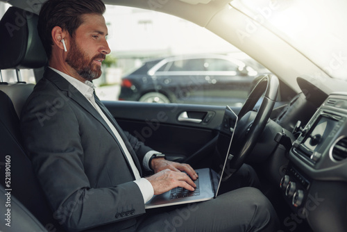 Handsome businessman working on laptop while sitting on driver seat in luxury car © Kostiantyn
