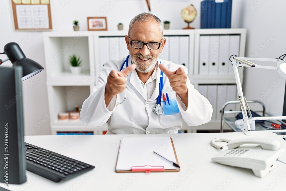 Mature doctor man at the clinic pointing fingers to camera with happy and funny face. good energy and vibes.