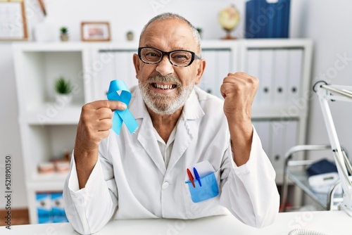 Mature doctor man holding blue ribbon at the clinic screaming proud, celebrating victory and success very excited with raised arms © Krakenimages.com