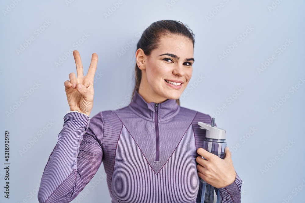 Beautiful woman wearing sportswear holding water bottle smiling looking to the camera showing fingers doing victory sign. number two. Beautiful woman wearing sportswear holding water bottle smiling looking to the camera showing fingers doing victory sign. number two.