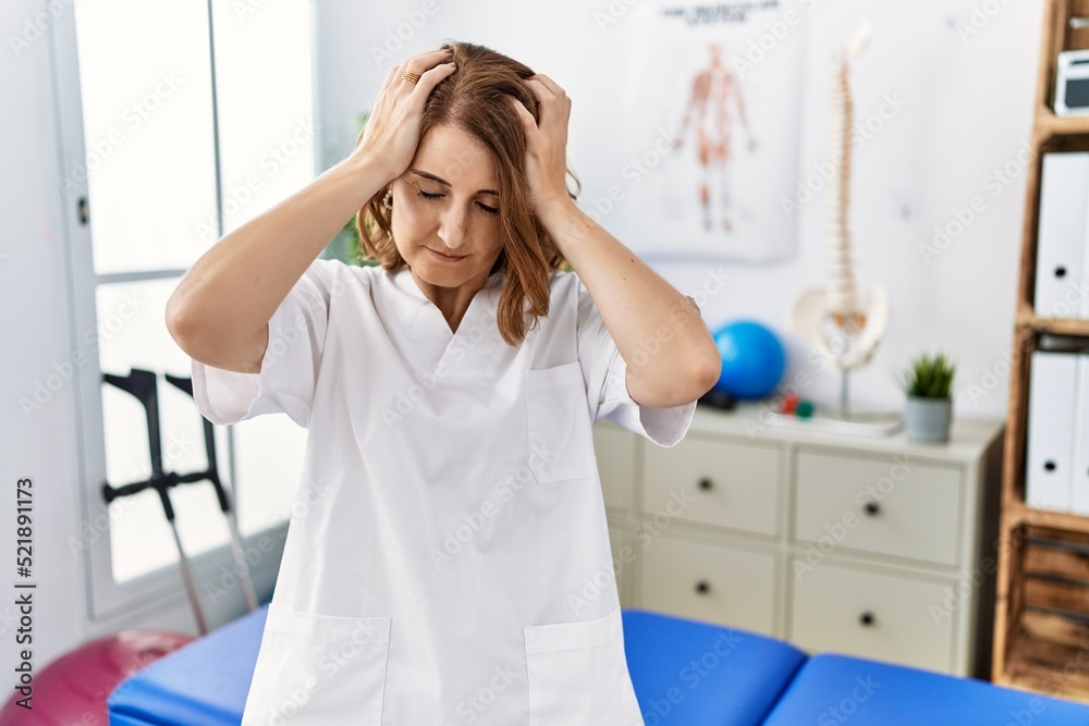 Middle age physiotherapist woman working at pain recovery clinic suffering from headache desperate and stressed because pain and migraine. hands on head. Middle age physiotherapist woman working at pain recovery clinic suffering from headache desperate and stressed because pain and migraine. hands on head.