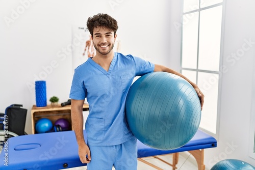 Hispanic physiotherapy man working at pain recovery clinic looking positive and happy standing and smiling with a confident smile showing teeth