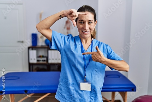Young hispanic woman wearing physiotherapist uniform standing at clinic smiling making frame with hands and fingers with happy face. creativity and photography concept.