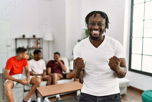 Young african man with friends at the living room very happy and excited doing winner gesture with arms raised  smiling and screaming for success. celebration concept.