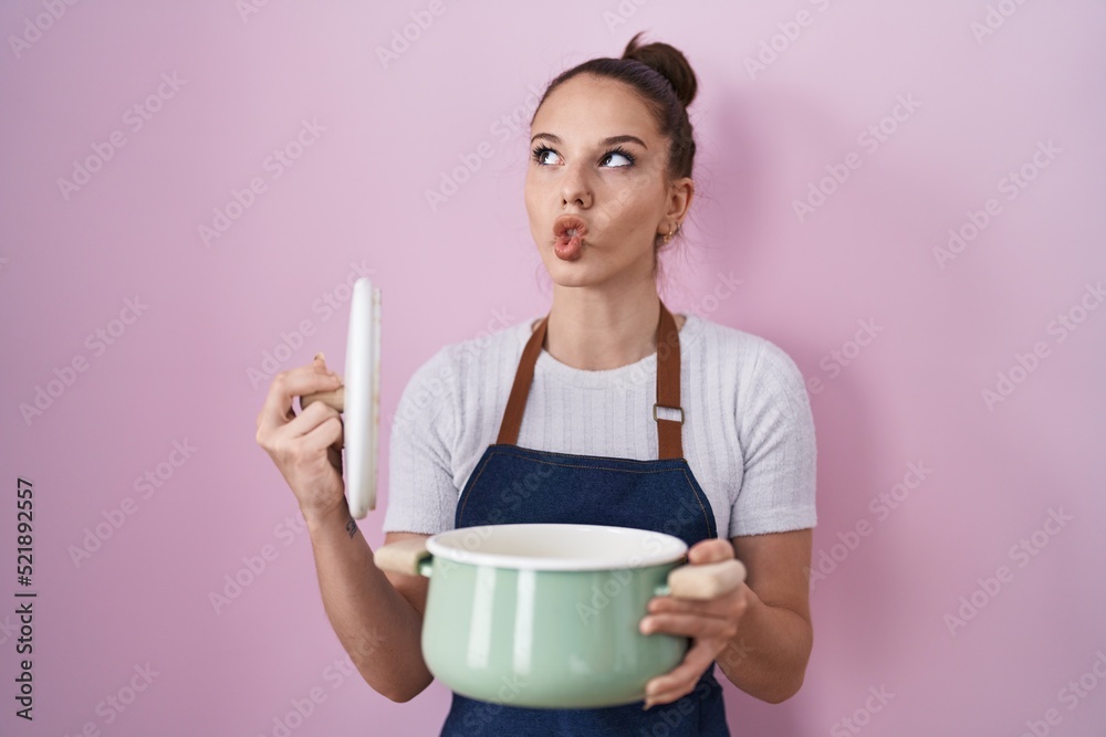Young hispanic girl wearing apron holding cooking pot making fish face with mouth and squinting eyes, crazy and comical. Young hispanic girl wearing apron holding cooking pot making fish face with mouth and squinting eyes, crazy and comical.
