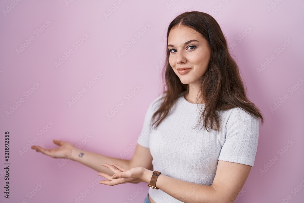 Young hispanic girl standing over pink background inviting to enter smiling natural with open hand Young hispanic girl standing over pink background inviting to enter smiling natural with open hand