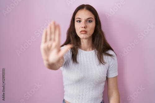 Young hispanic girl standing over pink background doing stop sing with palm of the hand. warning expression with negative and serious gesture on the face.