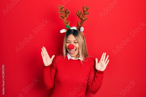 Beautiful hispanic woman wearing deer christmas hat and red nose celebrating mad and crazy for success with arms raised and closed eyes screaming excited. winner concept
