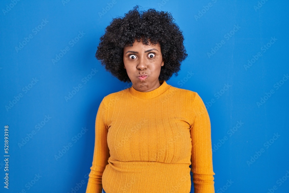Black woman with curly hair standing over blue background puffing cheeks with funny face. mouth inflated with air, crazy expression. Black woman with curly hair standing over blue background puffing cheeks with funny face. mouth inflated with air, crazy expression.