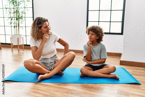 Young woman and son sitting on training mat at the gym serious face thinking about question with hand on chin, thoughtful about confusing idea