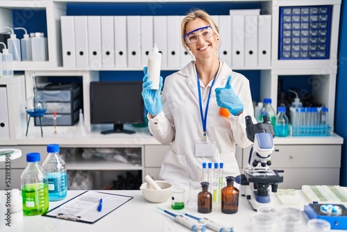 Young caucasian woman working at scientist laboratory holding body lotion smiling happy and positive  thumb up doing excellent and approval sign