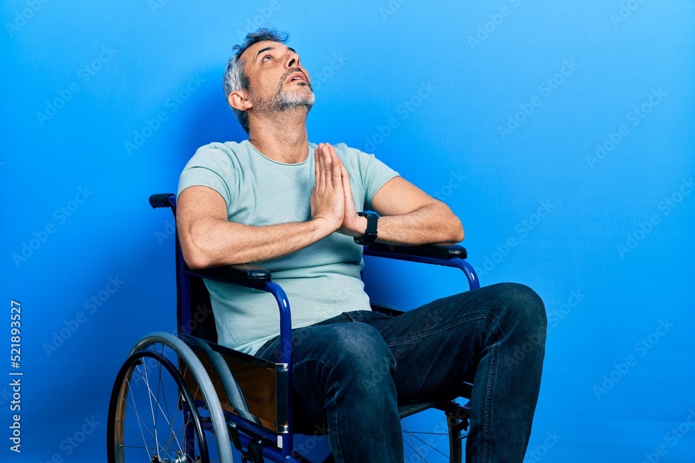 Fototapeta premium Handsome middle age man with grey hair sitting on wheelchair begging and praying with hands together with hope expression on face very emotional and worried. begging.
