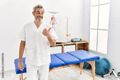 Middle age hispanic therapist man working at pain recovery clinic smiling with happy face looking and pointing to the side with thumb up.