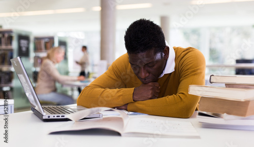 Thoughtful african-american man working with textbook and laptop in library. High quality photo