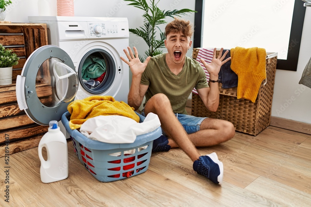 Young caucasian man putting dirty laundry into washing machine celebrating mad and crazy for success with arms raised and closed eyes screaming excited. winner concept Young caucasian man putting dirty laundry into washing machine celebrating mad and crazy for success with arms raised and closed eyes screaming excited. winner concept