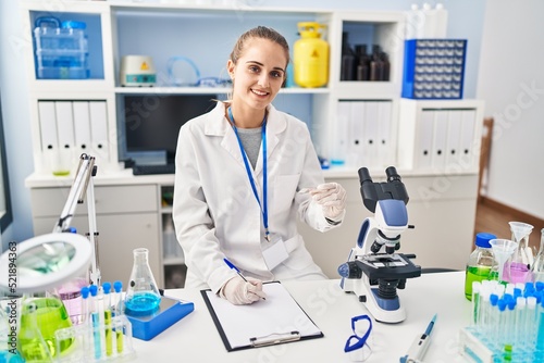Young blonde woman wearing scientist uniform writing on checklist at laboratory