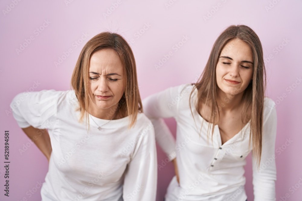 Middle age mother and young daughter standing over pink background suffering of backache, touching back with hand, muscular pain Middle age mother and young daughter standing over pink background suffering of backache, touching back with hand, muscular pain