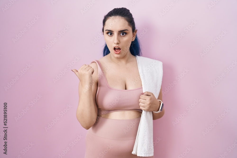 Young modern girl with blue hair wearing sportswear over pink background surprised pointing with hand finger to the side, open mouth amazed expression.