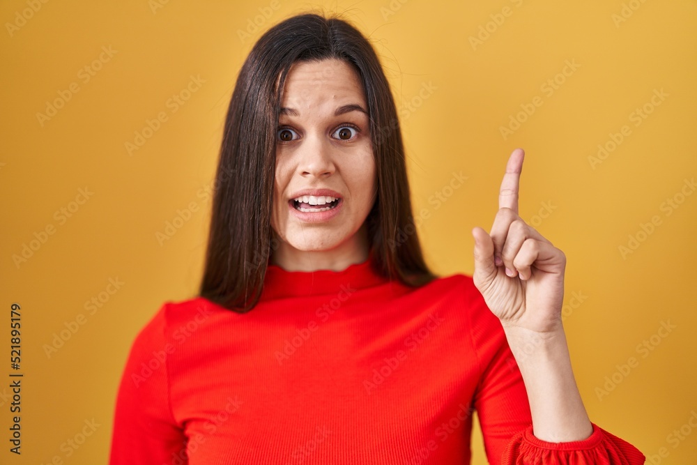 Young hispanic woman standing over yellow background pointing finger up with successful idea. exited and happy. number one. Young hispanic woman standing over yellow background pointing finger up with successful idea. exited and happy. number one.
