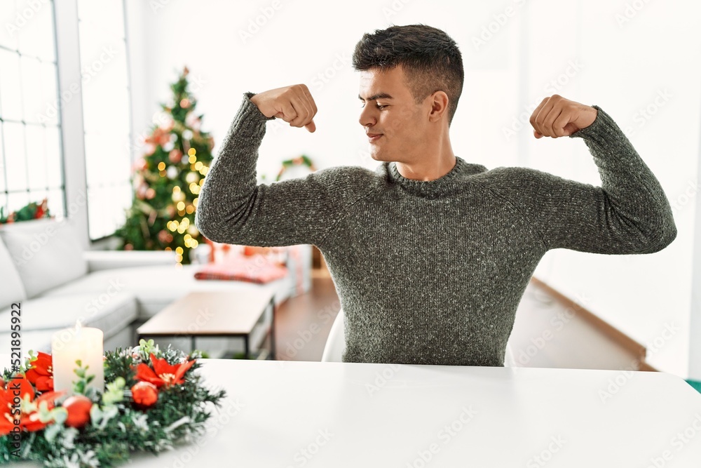Young hispanic man sitting on the table by christmas tree showing arms muscles smiling proud. fitness concept. Young hispanic man sitting on the table by christmas tree showing arms muscles smiling proud. fitness concept.