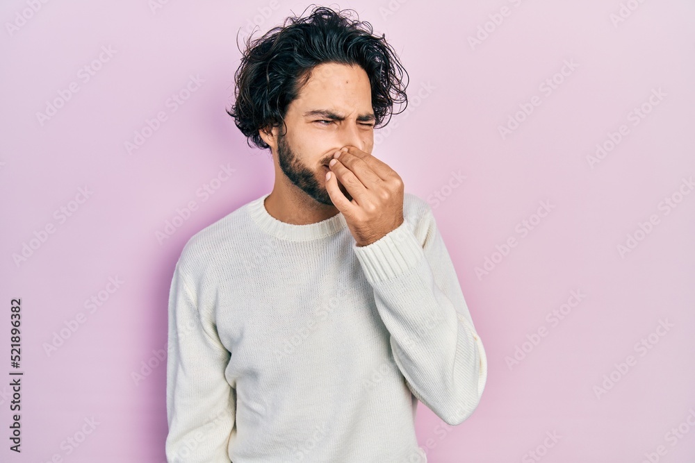 Handsome hispanic man wearing casual white sweater smelling something stinky and disgusting, intolerable smell, holding breath with fingers on nose. bad smell Handsome hispanic man wearing casual white sweater smelling something stinky and disgusting, intolerable smell, holding breath with fingers on nose. bad smell