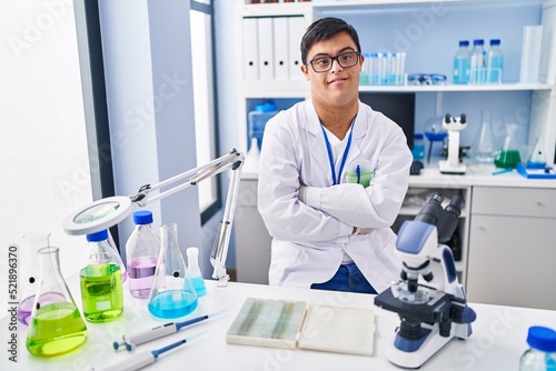 Down syndrome man wearing scientist uniform sitting with arms crossed gesture at laboratory