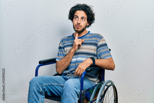 Handsome hispanic man sitting on wheelchair thinking concentrated about doubt with finger on chin and looking up wondering