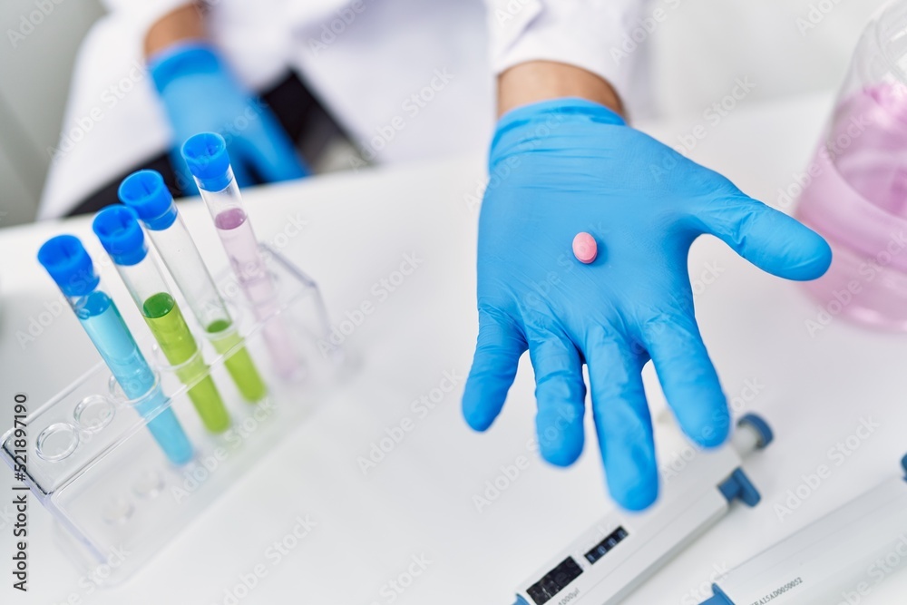 Young hispanic man wearing scientist uniform holding pill at laboratory