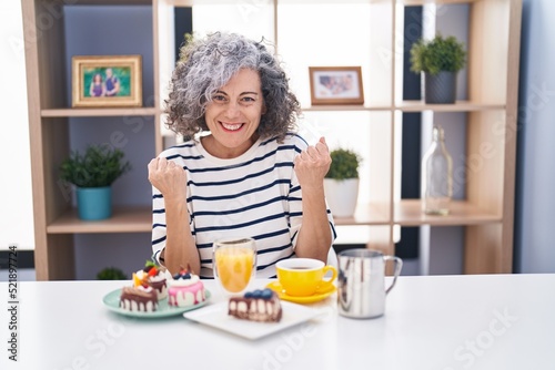 Middle age woman with grey hair eating pastries and drinking coffee for breakfast very happy and excited doing winner gesture with arms raised, smiling and screaming for success. celebration concept.