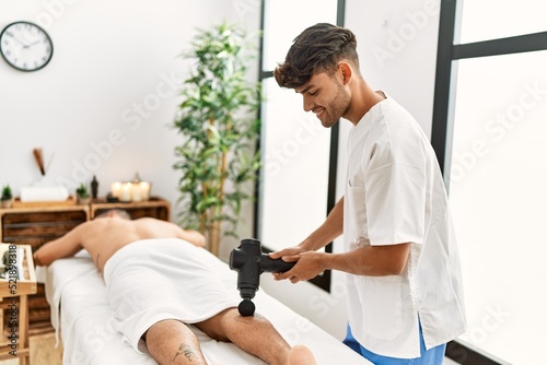 Two hispanic men physiotherapist and patient massaging legs using percussion gun at beauty center