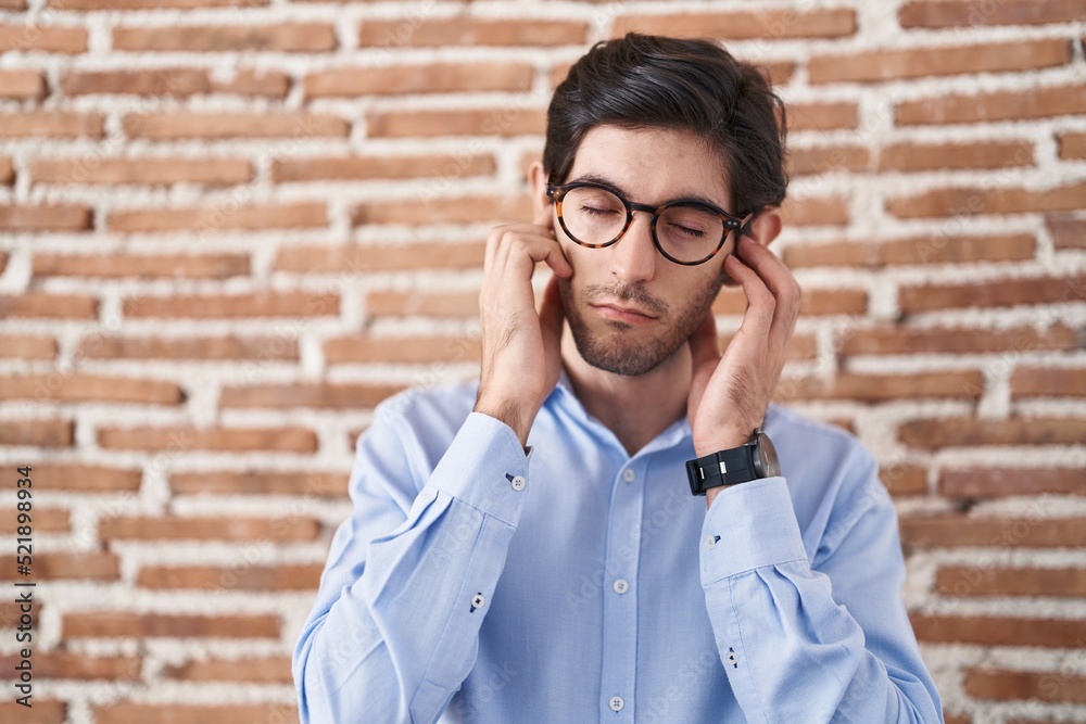 Young hispanic man standing over brick wall background covering ears with fingers with annoyed expression for the noise of loud music. deaf concept. Young hispanic man standing over brick wall background covering ears with fingers with annoyed expression for the noise of loud music. deaf concept.
