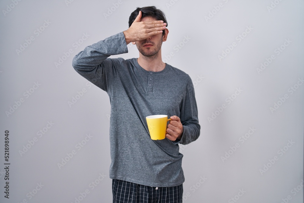 Young hispanic man wearing pajama drinking a cup of coffee covering eyes with hand, looking serious and sad. sightless, hiding and rejection concept Young hispanic man wearing pajama drinking a cup of coffee covering eyes with hand, looking serious and sad. sightless, hiding and rejection concept