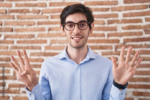 Young hispanic man standing over brick wall background showing and pointing up with fingers number nine while smiling confident and happy.