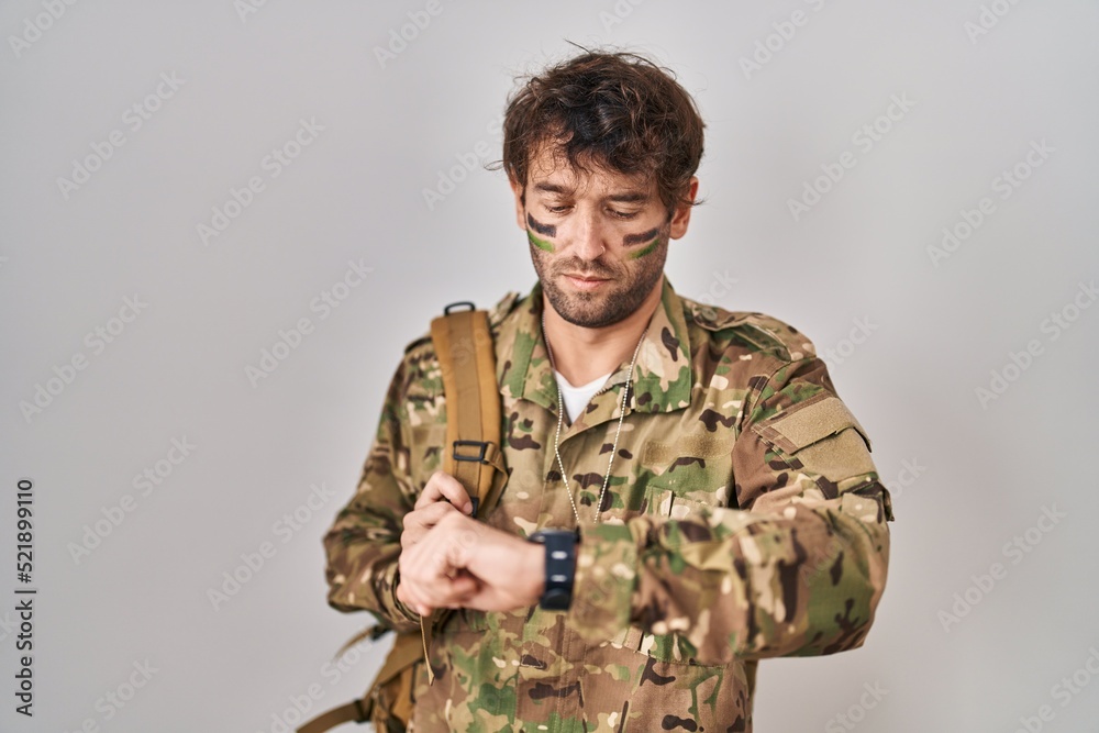 Hispanic young man wearing camouflage army uniform checking the time on wrist watch, relaxed and confident