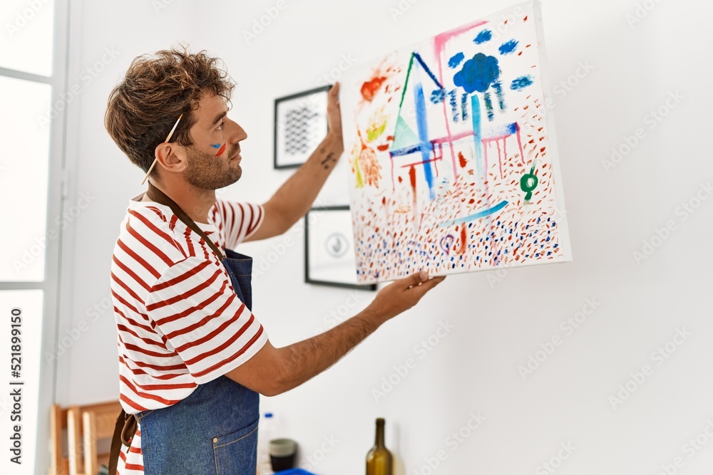Young hispanic man hanging canvas on wall at art studio Young hispanic man hanging canvas on wall at art studio
