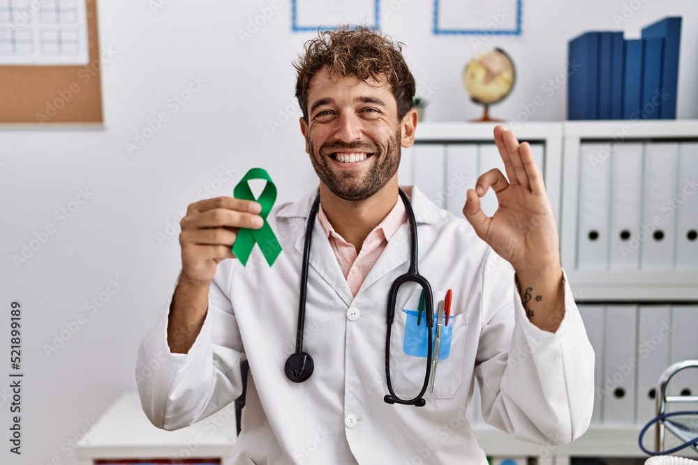 Young hispanic doctor man holding support green ribbon at clinic doing ok sign with fingers, smiling friendly gesturing excellent symbol Young hispanic doctor man holding support green ribbon at clinic doing ok sign with fingers, smiling friendly gesturing excellent symbol