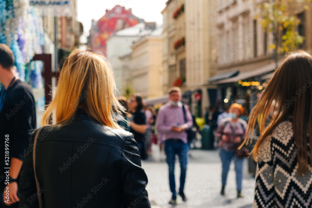 Defocus two stylish blonde and white women walking outdoors in autumn city street at sunset time wearing black jacket. View from the back. Friendship and travel. Out of focus
