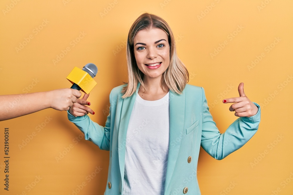 Beautiful caucasian woman being interviewed by reporter holding microphone looking confident with smile on face, pointing oneself with fingers proud and happy. Beautiful caucasian woman being interviewed by reporter holding microphone looking confident with smile on face, pointing oneself with fingers proud and happy.