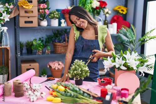 Young hispanic woman florist talking on smartphone cutting plant at florist store