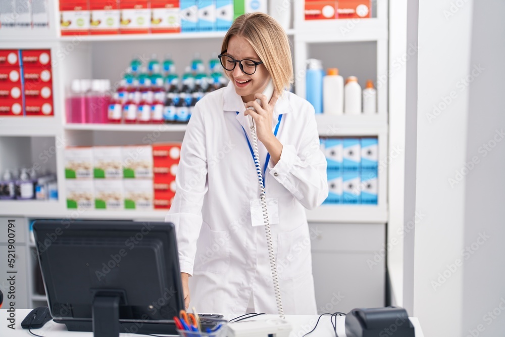 Young blonde woman pharmacist talking on telephone using computer at pharmacy
