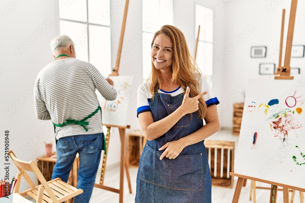 Hispanic woman wearing apron at art studio cheerful with a smile of face pointing with hand and finger up to the side with happy and natural expression on face