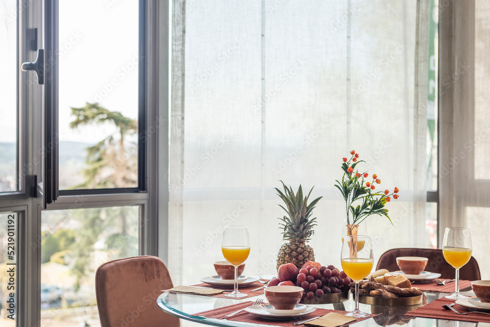 Fototapeta premium Dining room with a circular glass table full of assorted fruits, glasses of orange juice next to a gazebo with a view